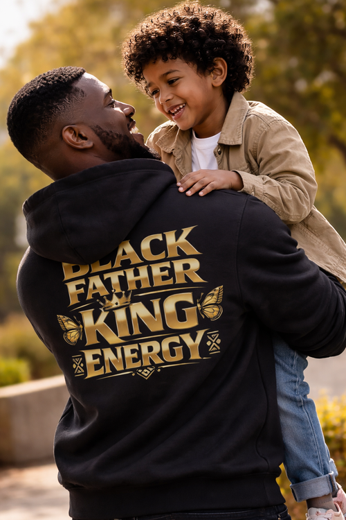 Man wearing a hoodie with 'Black Father King Energy' text, holding a child outdoors.